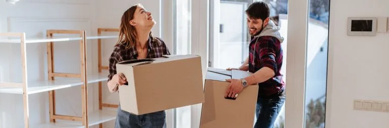 A young couple carrying moving boxes into their first home, looking excited.
